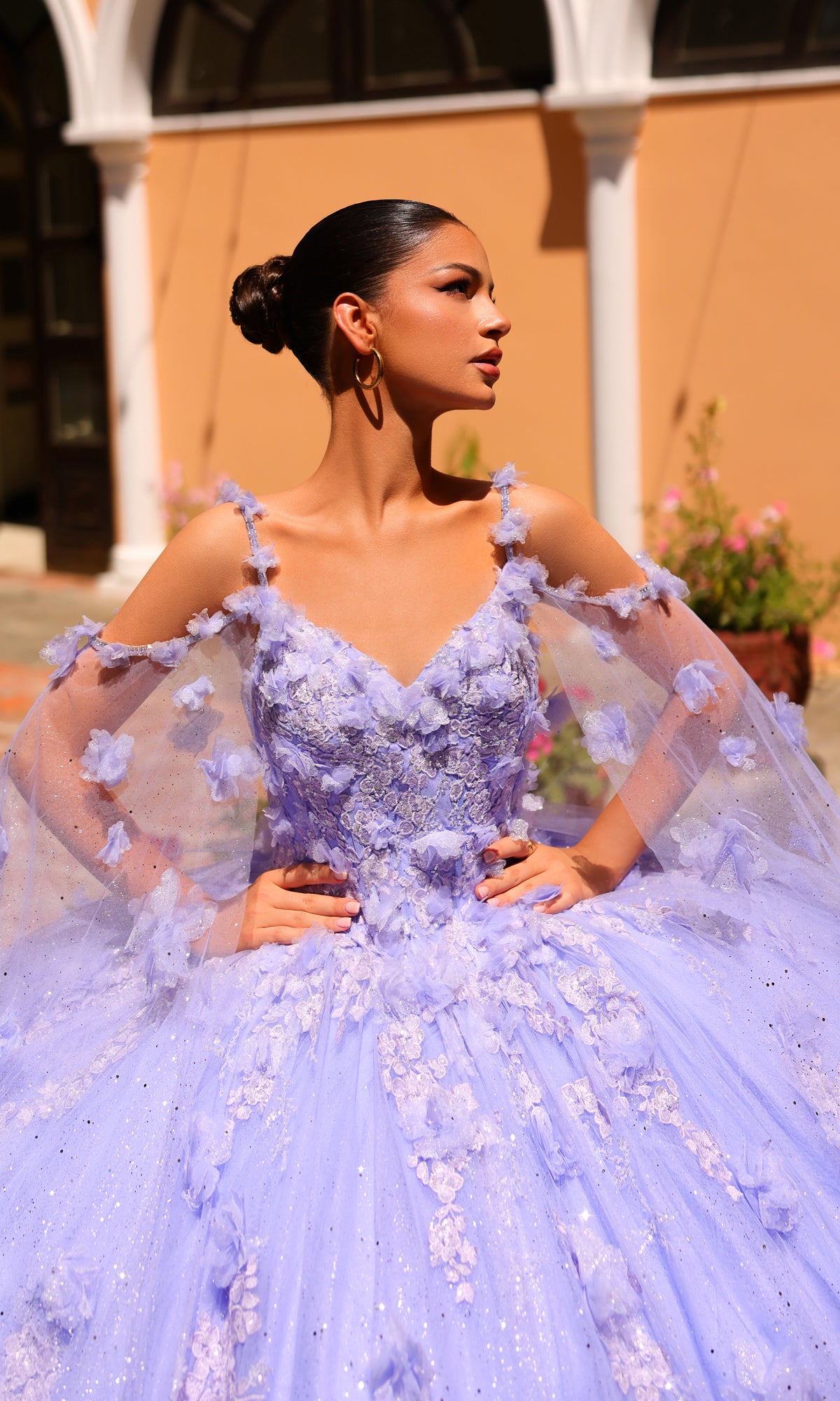 A woman poses outdoors in the Amarra 54254 Periwinkle Purple Quincea¤era Dress featuring floral appliqu‚s and tulle, her hair in a bun, hands on hips, gazing to the side.